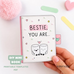 Könnte beinhalten: Ein weißes DIY-Minibuch mit rosa Buchrücken und einem Cover, das zwei lächelnde Marshmallow-Figuren, kleine Herzen und Sterne zeigt. Der Text lautet "BESTIE, YOU ARE..." mit einer kleinen rosa Lasche an der Seite, die ein grünes Herz zeigt. Das Design ist mit winzigen bunten Perlen verziert.