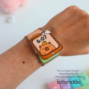 May include: A paper craft watch with a cartoon bear face on the screen, displaying the time 6:07. The watch band is tan with a cloud design. The text "katemade" is visible.