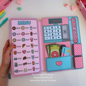 May include: A pink and blue play cash register with a menu of donuts and drinks. The register has a keypad, a card reader, and a drawer with play money and a credit card. The menu lists various donuts, pastries, and beverages.