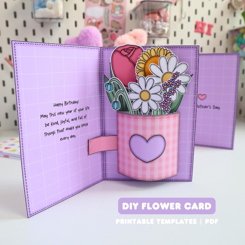 May include: A handmade, pop-up greeting card with a floral arrangement. The card is lavender with a grid pattern and features a pink gingham vase with a heart. The flowers include a pink rose, daisy, and sunflower. Text reads "Happy Birthday!" and "DIY FLOWER CARD".