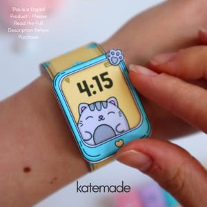 May include: A paper-based smartwatch with a light blue frame and a yellow screen displaying the time 4:15. The screen features a cartoon cat illustration. The watch band is yellow with blue cloud designs. The text "katemade" is at the bottom.