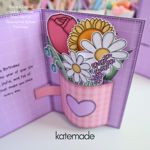 May include: A handmade greeting card with a pop-up floral arrangement. The card features a pink gingham mug with a heart, holding a pink rose, sunflower, daisies, and purple flowers. The card is lavender with the text "katemade".