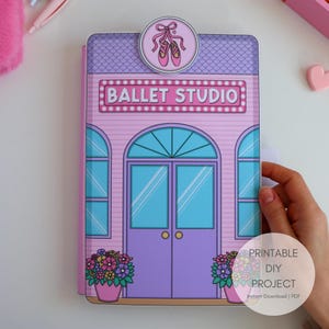 Puede incluir: Un proyecto de bricolaje imprimible de Ballet Studio rosa y morado. El edificio tiene una puerta, ventanas y un letrero que dice "BALLET STUDIO". Un pin de zapatilla de ballet está en la parte superior. El edificio tiene macetas.