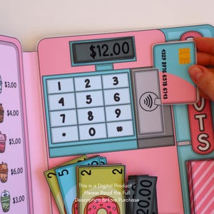 May include: A pink and blue play cash register with a keypad, a digital display showing $12.00, and a credit card. The register has a slot for contactless payments. Several play money bills and cards are also visible.