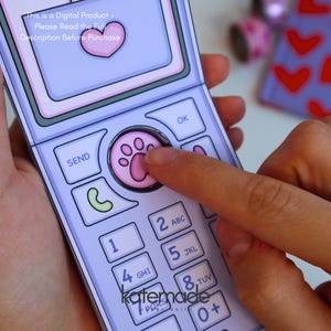 May include: A light purple paper flip phone with a pink paw print button, a screen displaying a pink heart, and a standard numeric keypad. The text on the screen reads: This is a Digital Product - Please Read the Full Description Before Purchase.