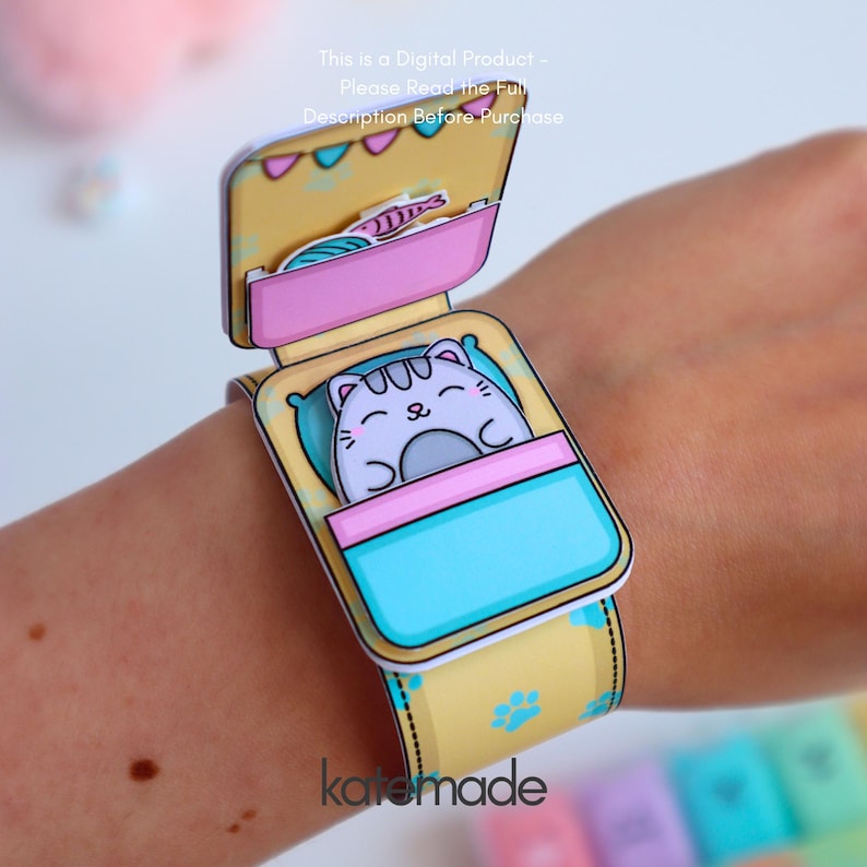 May include: A papercraft cat-themed bracelet with a sleeping cat illustration. The bracelet is yellow with blue paw prints and a pink and blue bed design. The text "katemade" is visible.