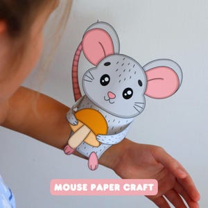 Puede incluir: Un juguete de papel con forma de ratón gris con grandes orejas forradas de rosa, ojos negros y una pequeña nariz rosa. El ratón sostiene un hongo amarillo y beige. El texto MOUSE PAPER CRAFT está impreso en blanco sobre un fondo rosa.