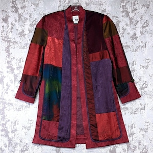 May include: A long-sleeved jacket featuring a patchwork design of crinkled fabric in shades of red, purple, and dark blue. The garment has an open front with a vertical purple trim, two large front pockets, and a label at the inner neckline.