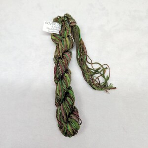 May include: A skein of hand-dyed yarn in shades of olive green, brown, and metallic copper. The yarn features a variety of textures, including ribbon-like strands and thin metallic threads. A white tag labeled Oliver Twists is attached.