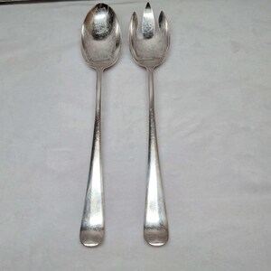 May include: A pair of silver-toned metal salad serving utensils consisting of one large solid spoon and one large three-pronged fork. Both pieces feature long, smooth, tapered handles and a polished, reflective metallic finish throughout.