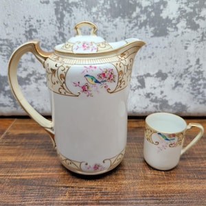 May include: A porcelain teapot with a lid and a matching small cup, both featuring a white base, gold trim, and a delicate floral pattern with a small blue bird perched on a branch, accented by intricate gold scrollwork around the rims.