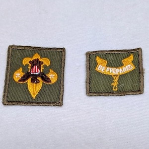 May include: Two square olive green embroidered patches. The left patch features a yellow fleur-de-lis emblem with a central eagle and red, white, and blue shield. The right patch displays a yellow ribbon banner with the text BE PREPARED.