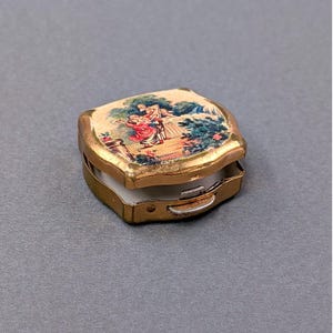 May include: A small, antique, gold-toned metal pillbox with a hinged lid. The lid features a colorful, detailed illustration of figures in a garden setting. The interior is white. The pillbox is open, revealing the inside.