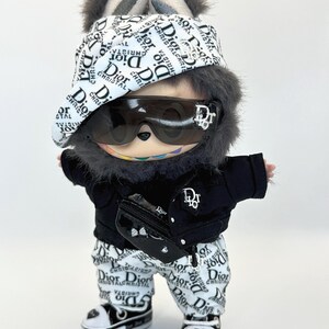 May include: A plush toy dressed in a black jacket, white pants, and sneakers, all adorned with the "Dior" logo. The toy wears sunglasses, a hat, and a fanny pack, also branded with the same logo. The toy has a furry head with bunny ears.