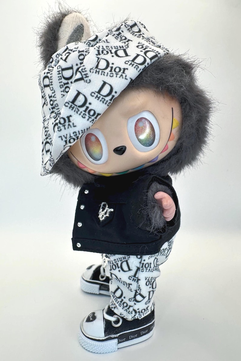 May include: A collectible figurine wearing a black jacket, pants, and shoes, all adorned with the "Dior" logo in white on a black background. The figurine has a furry hood and hat, and its eyes have a rainbow effect.