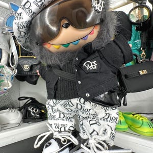 May include: A doll wearing a black jacket, white pants, and a beret, all with a Dior logo pattern. The doll has a pair of black and white sneakers and a small black purse. The doll is wearing sunglasses.