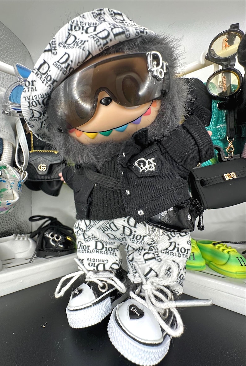 May include: A doll wearing a black jacket, white pants, and a beret, all with a Dior logo pattern. The doll has a pair of black and white sneakers and a small black purse. The doll is wearing sunglasses.