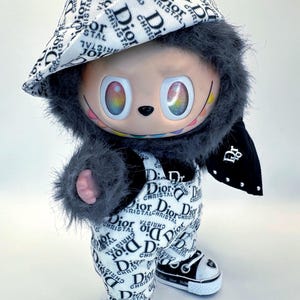 May include: A plush doll wearing a hat, overalls, and sneakers, all featuring a black and white "Dior" logo pattern. The doll has a gray furry hood and arms, and rainbow-colored eyes.