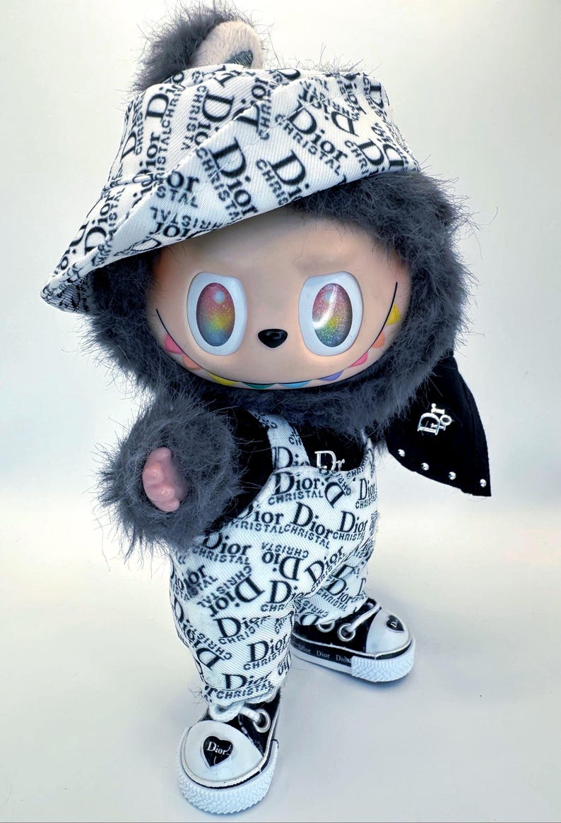 May include: A plush doll wearing a hat, overalls, and sneakers, all featuring a black and white "Dior" logo pattern. The doll has a gray furry hood and arms, and rainbow-colored eyes.