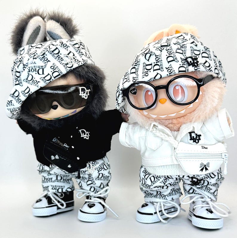 May include: Two plush figures dressed in matching outfits. One figure wears a black jacket, sunglasses, and a hat with a fur trim. The other figure wears a white jacket and glasses. Both figures have matching pants, hats, and shoes with the Dior logo.