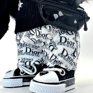 May include: Close-up of a doll wearing a black and white outfit. The doll has on white pants with a Dior logo print, black and white sneakers, a black fanny pack, and a black sweater. A Dior logo charm hangs from the pants.