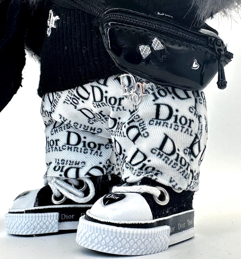 May include: Close-up of a doll wearing a black and white outfit. The doll has on white pants with a Dior logo print, black and white sneakers, a black fanny pack, and a black sweater. A Dior logo charm hangs from the pants.
