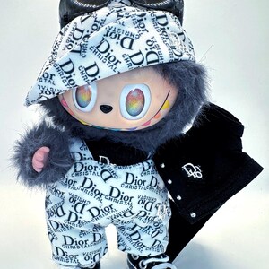 May include: A plush doll wearing a white and black patterned outfit with the word "Dior" repeated. The doll has a black jacket, sunglasses, and sneakers. The doll's eyes are rainbow-colored.
