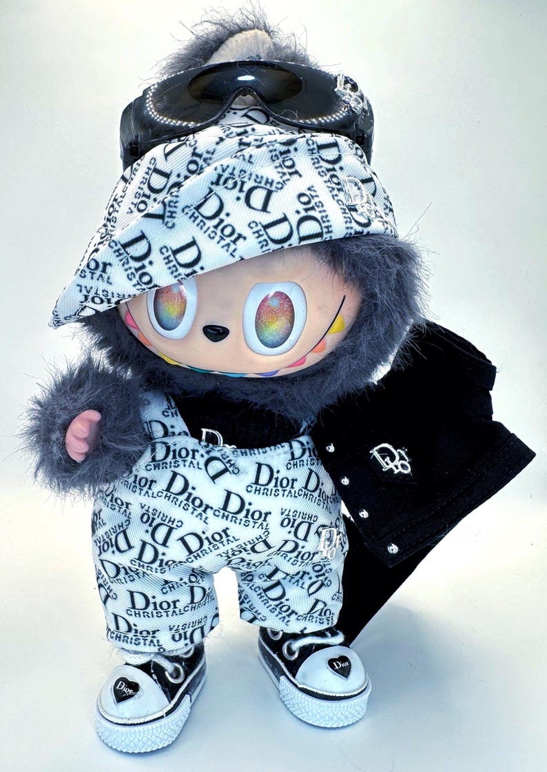 May include: A plush doll wearing a white and black patterned outfit with the word "Dior" repeated. The doll has a black jacket, sunglasses, and sneakers. The doll's eyes are rainbow-colored.