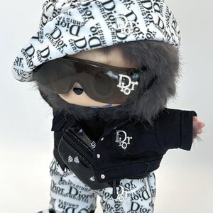 May include: A doll dressed in a Dior-branded outfit, including a hat, sunglasses, jacket, pants, and shoes. The outfit is primarily black and white with the Dior logo. The doll has gray fur and bunny ears.