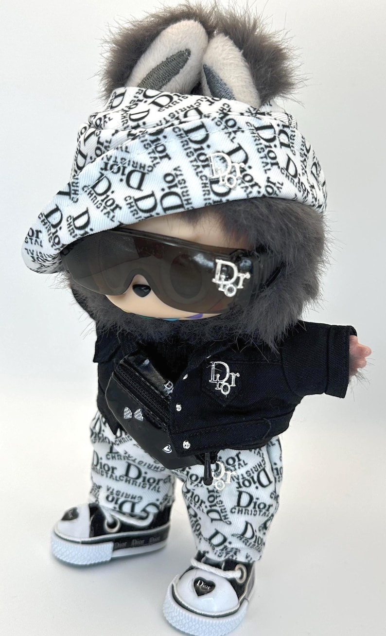 May include: A doll dressed in a Dior-branded outfit, including a hat, sunglasses, jacket, pants, and shoes. The outfit is primarily black and white with the Dior logo. The doll has gray fur and bunny ears.