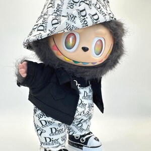 May include: A whimsical figurine dressed in a black jacket, pants, and sneakers, all adorned with the "Dior" logo. The figure has a colorful face, a furry head, and a matching hat. The sneakers have white soles.