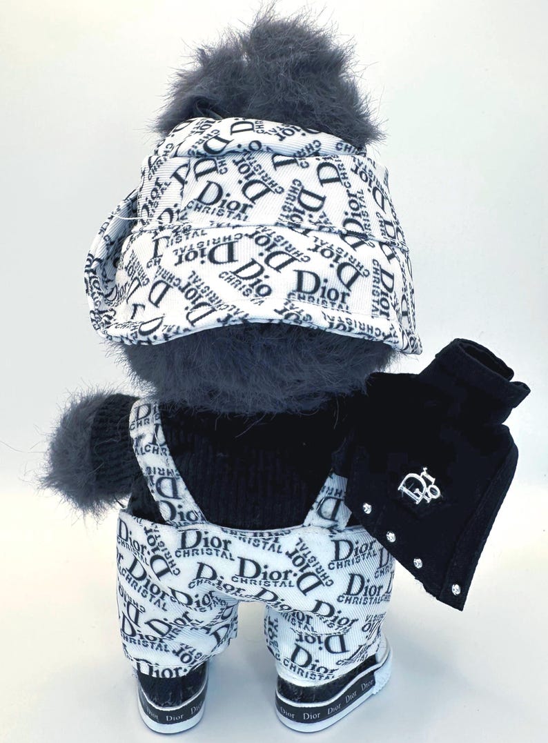 May include: A plush toy dressed in a black and white outfit with the "Dior" logo. The outfit includes a hat, overalls, jacket, and sneakers. The jacket has a "Dior" logo and rhinestone accents.