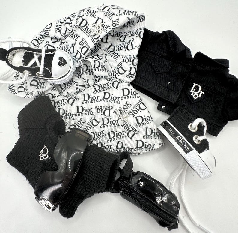 May include: A collection of miniature fashion items, including a black jacket, sneakers, and a small purse. The items feature the "Dior" logo in black and white. The sneakers have white laces and a heart design.