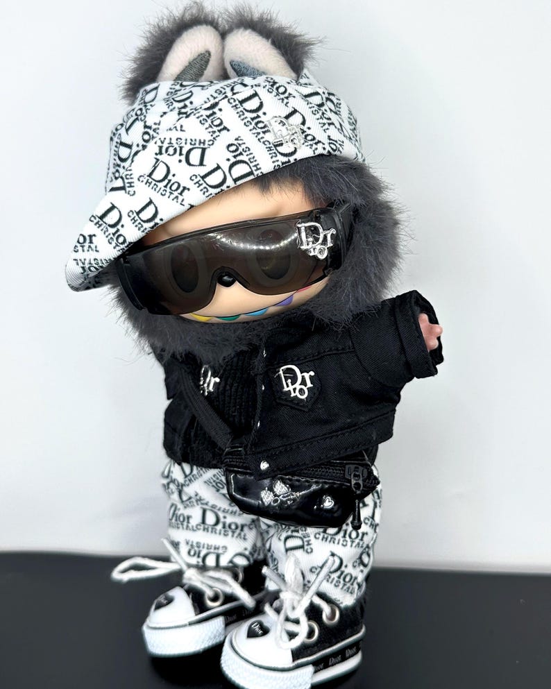 May include: A doll wearing a black jacket, white and black pants, and black and white sneakers. The doll is also wearing sunglasses and a hat with the text "Dior" printed on it. The doll has a small black purse.