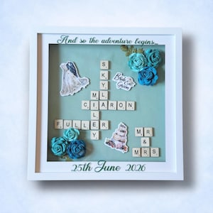 May include: A framed wedding keepsake with a teal background. The frame includes paper flowers, a wedding dress graphic, and Scrabble tiles spelling out names. The text "And so the adventure begins..." and "25th June 2026" are also visible.