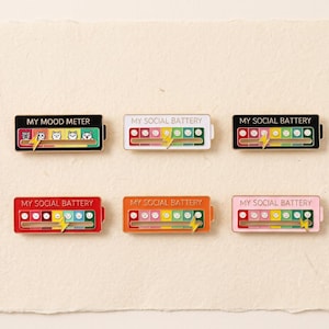 May include: Six rectangular enamel pins shaped like batteries. One reads MY MOOD METER with cat faces, while five read MY SOCIAL BATTERY with emoji faces ranging from sad to happy. Each pin features a sliding yellow lightning bolt indicator.