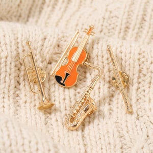Peut inclure: Un ensemble de quatre broches en métal doré représentant des instruments de musique : une trompette, un violon orange et noir avec son archet, un saxophone et une flûte. Chaque broche présente une finition métallique polie.