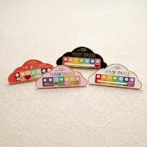 Puede incluir: Cuatro pines de esmalte con forma de nube y la inscripción "my MOOD TRACKER" en negro, rojo, rosa y blanco. Cada pin presenta una escala horizontal de arcoíris con siete expresiones faciales y un indicador deslizante en forma de corazón.