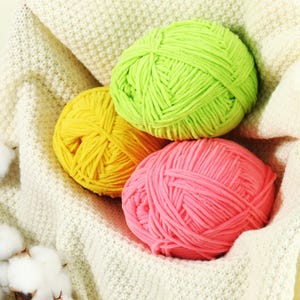 May include: Three balls of brightly coloured yarn: yellow, pink, and green. The yarn sits on a white knitted blanket. The yarn is soft and fluffy, ideal for knitting or crochet projects.