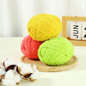 May include: Three balls of yarn in bright colors: yellow, coral, and lime green. The yarn balls are stacked on a small wooden plate. Cotton bolls are in the foreground. A wooden calendar shows the month of June.