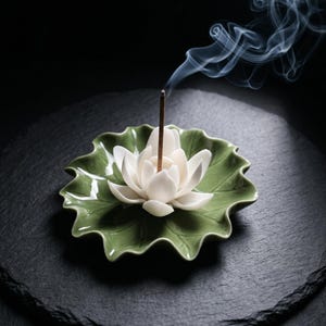 May include: A ceramic incense holder shaped like a green lily pad with a white lotus flower in the center. A thin brown incense stick is inserted into the middle of the lotus, emitting a thin trail of gray smoke that curls upward.