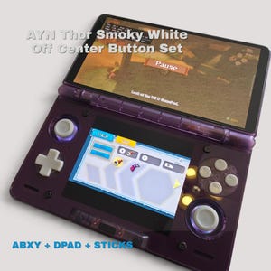 May include: A handheld gaming console with a translucent purple shell, featuring a white D-pad, white ABXY buttons, and white analog sticks. The screen displays a game menu with the text AYN Thor Smoky White Off Center Button Set.