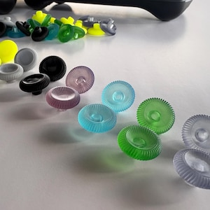May include: A collection of colorful, translucent controller thumbsticks in various colors, including yellow, gray, black, purple, blue, and green. The thumbsticks have a ridged design and are arranged on a white surface.