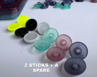 May include: A collection of circular, textured joystick caps in various translucent and solid colors including neon yellow, black, white, teal, pink, and purple. The text 2 STICKS + A SPARE is printed in white on the surface near the joystick caps.