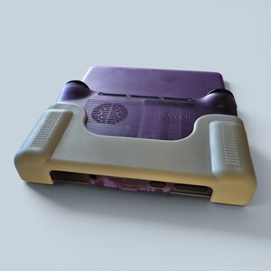 Puede incluir: Una consola de juegos port&aacute;til con carcasa morada transl&uacute;cida y funda protectora de silicona beige. El dispositivo presenta una rejilla de ventilaci&oacute;n circular en la parte posterior, el logotipo AYN y zonas de agarre texturizadas en los laterales.