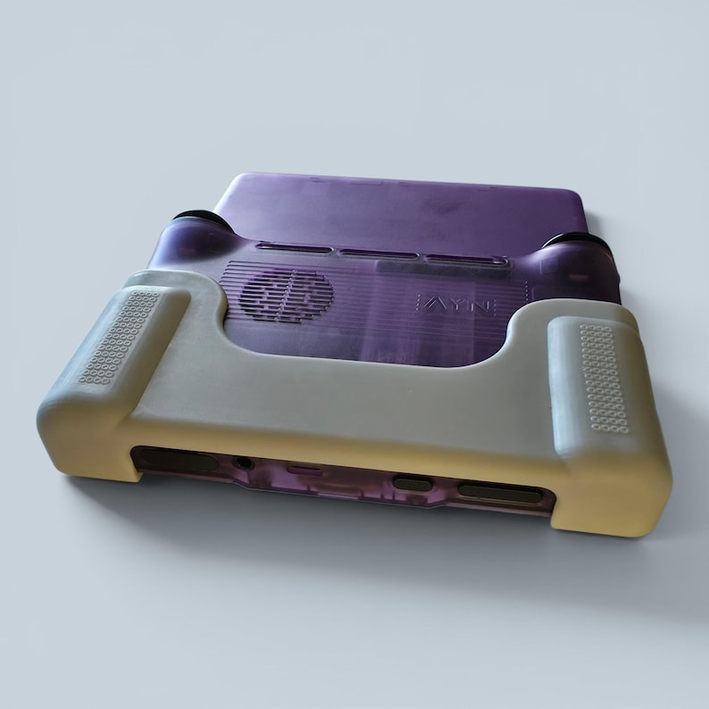 Puede incluir: Una consola de juegos port&aacute;til con carcasa morada transl&uacute;cida y funda protectora de silicona beige. El dispositivo presenta una rejilla de ventilaci&oacute;n circular en la parte posterior, el logotipo AYN y zonas de agarre texturizadas en los laterales.