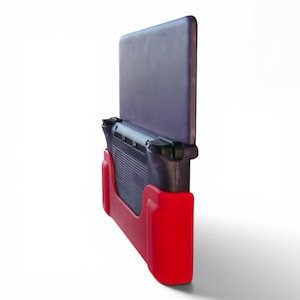 Puede incluir: Una consola de juegos port&aacute;til con una carcasa de color morado oscuro transl&uacute;cido y una funda protectora de color rojo vibrante. El dispositivo se muestra desde un &aacute;ngulo lateral, destacando los botones superiores, las rejillas de ventilaci&oacute;n y la funda.