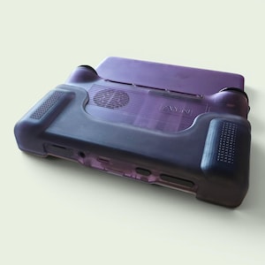 Puede incluir: Una funda de consola de juegos port&aacute;til de color morado transl&uacute;cido con agarre texturizado en los laterales, un recorte circular para el ventilador en la parte posterior y el texto AYN en relieve visible en la superficie trasera del dispositivo.