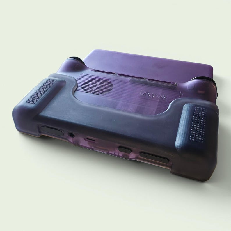 Puede incluir: Una funda de consola de juegos port&aacute;til de color morado transl&uacute;cido con agarre texturizado en los laterales, un recorte circular para el ventilador en la parte posterior y el texto AYN en relieve visible en la superficie trasera del dispositivo.