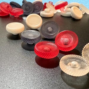 May include: A collection of various knobs in clear, red, black, and cream colors. The knobs have different shapes and designs, including round, ribbed, and textured surfaces. Some knobs are on a black surface.